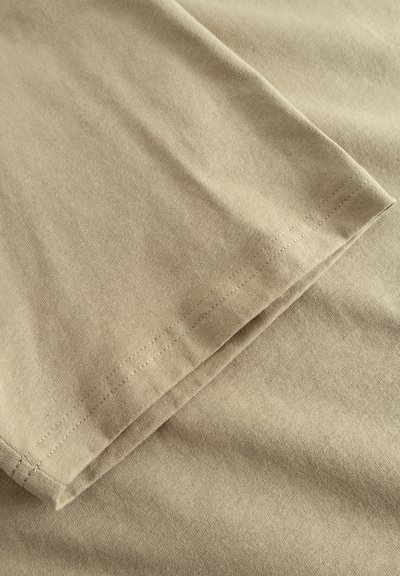 Tissu en coton beige avec une texture lisse, présentant un ourlet replié et des coutures visibles le long du bord. Pas de motifs ou d'accents notables.