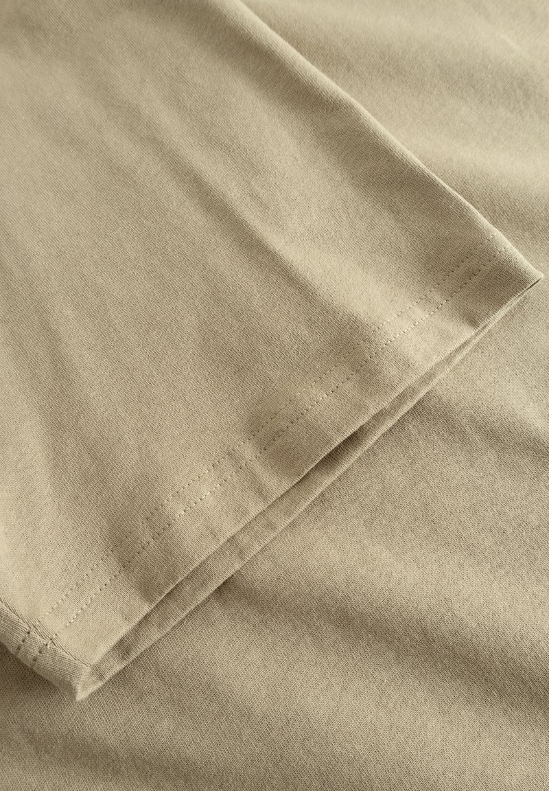 Tela de algodón beige con una textura suave, que presenta un dobladillo doblado y costuras visibles a lo largo del borde. Sin patrones o acentos notables.