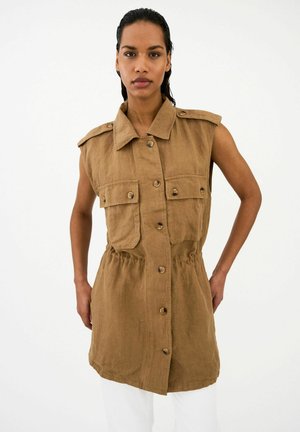 Femme portant une robe marron sans manches à boutons sur le devant avec poches, debout les mains sur les hanches sur un fond blanc uni.
