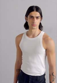 White ribbed tank top with a round neckline, worn by a person with long dark hair, standing against a neutral background.