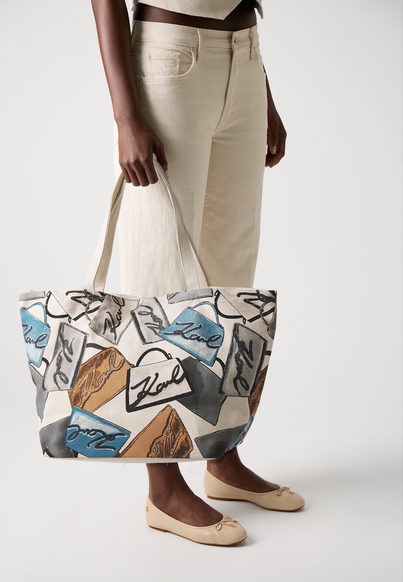 Personne portant un pantalon beige et des chaussures tenant un grand sac en toile avec des illustrations multicolores de sacs à main et le mot "Karl" écrit dessus.
