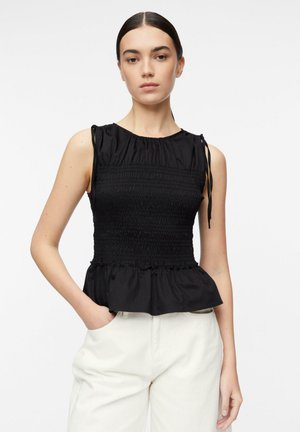 Giovane donna con capelli scuri raccolti in uno chignon basso, indossa un top nero senza maniche arricciato con orlo a peplo e pantaloni bianchi, in piedi davanti a uno sfondo bianco.