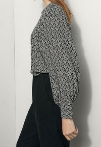 Schwarz-weiß gemusterte Bluse mit hohem Kragen, puffigen Ärmeln und lockerer Passform, kombiniert mit schwarzen Hosen. Glatte Stofftextur.
