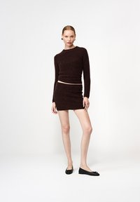 Hnedý nadýchaný sveter s dlhými rukávmi, v kombinácii s ladenou hnedou mini sukňou. Modelka má na sebe čierne ploché topánky. Jednoduchý, minimalistický dizajn.