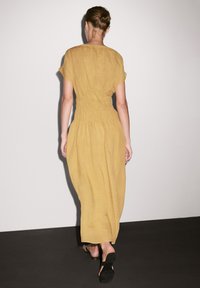 Senfgelbes Kleid mit kurzen Ärmeln, geraffter Taille und fließender Silhouette. Glatte Leinenstruktur, getragen mit schwarzen Riemchensandalen.