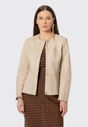 Mujer con cabello largo y castaño que lleva una chaqueta de cuero beige sobre un vestido marrón con lunares, complementada con un pendiente largo de oro en la oreja izquierda.