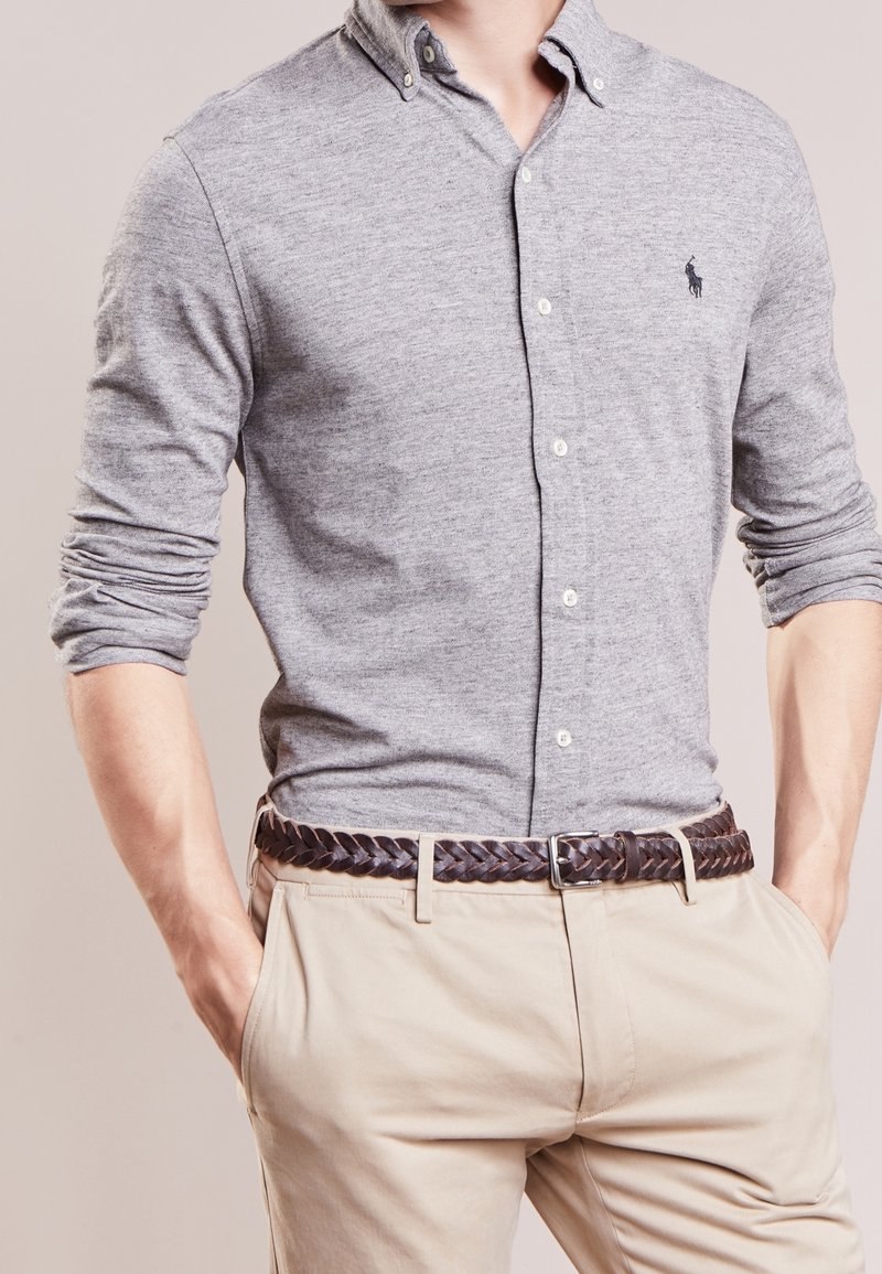 Szara koszula z długim rękawem, zapinana na guziki, z subtelną teksturą i logo konia, w zestawieniu z beżowymi spodniami i plecionym brązowym skórzanym paskiem.