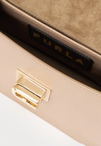 Εσωτερικό μπεζ δερμάτινης τσάντας με χρυσή μεταλλική αγκράφα, με σουέτ επένδυση και μαύρη ετικέτα που γράφει "FURLA" με χρυσά γράμματα.