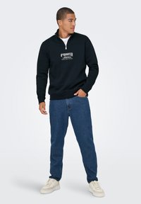 Zwarte hoodie met rits en een geprinte "4th Quarter Athletics" logo, gecombineerd met blauwe jeans en witte sneakers, tegen een lichte achtergrond.