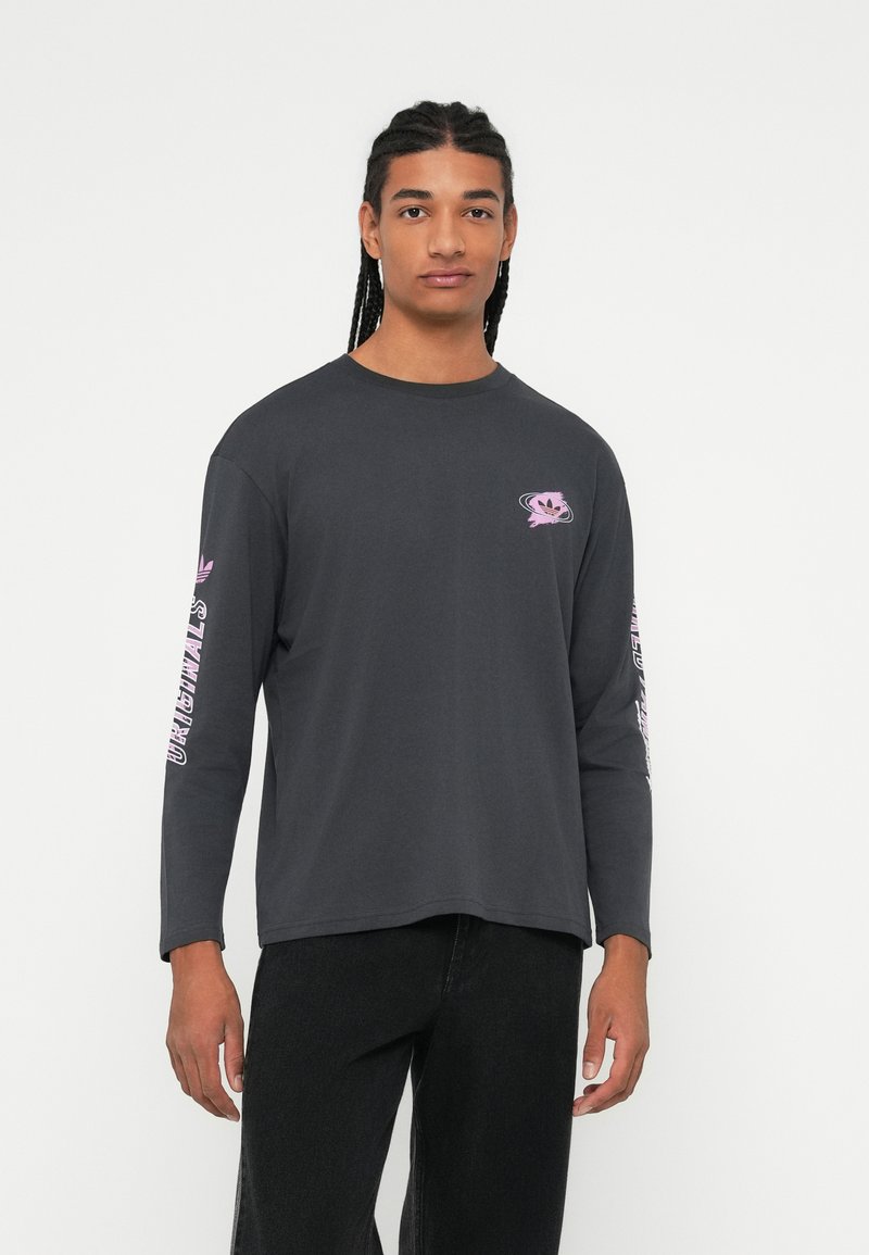 Junge Person mit geflochtenem Haar, die ein dunkles langärmliges Shirt mit pinkfarbenen Logos und schwarze Hosen trägt, steht vor einem schlichten weißen Hintergrund.