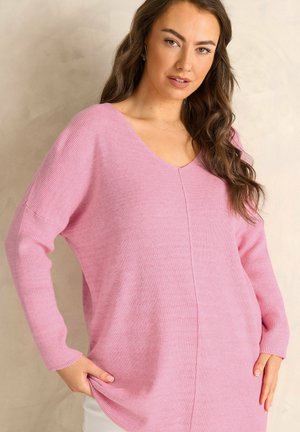 Jonge vrouw met lang golvend haar, gekleed in een losse roze trui met V-hals, staand met handen in de zakken tegen een neutrale achtergrond.