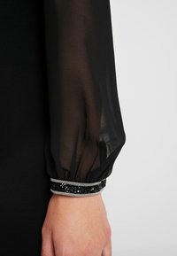 Durchsichtiges schwarzes Langarmshirt mit einem Ärmelabschluss, der mit kleinen schwarzen Perlen und silberner Verzierung dekoriert ist, getragen von einer Person mit heller Hautfarbe.