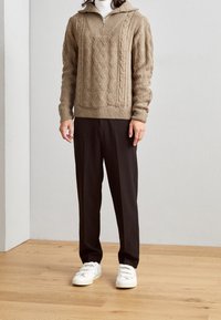 Beiger gestrickter Pullover mit Reißverschlusskragen, ausgestattet mit einem strukturierten Zopfmuster, kombiniert mit schwarzen Hosen und weißen Turnschuhen auf einem Holzboden.