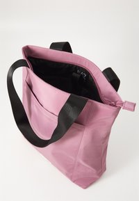 Rosa totebag med teksturert ytre, svarte stropper, glidelås lukking, og en frontlomme. Innvendig har den svart fôr.