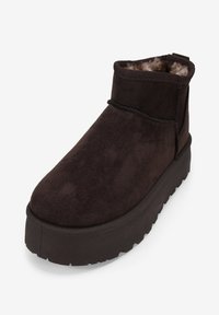 Botines de ante marrón con un interior suave, punta redonda y suela de goma gruesa que presenta un patrón texturizado. Diseño minimalista con detalles de costura.