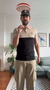 Camisa polo de manga corta con la parte superior beige y la parte inferior negra. Los pantalones son de estilo cargo en beige claro. El fondo presenta vegetación y un sofá.