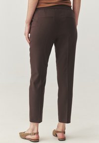 Person, der dunkle braune Slim-Fit-Hosen und beigefarbene flache Slingback-Schuhe trägt, steht vor einem hellen neutralen Hintergrund.