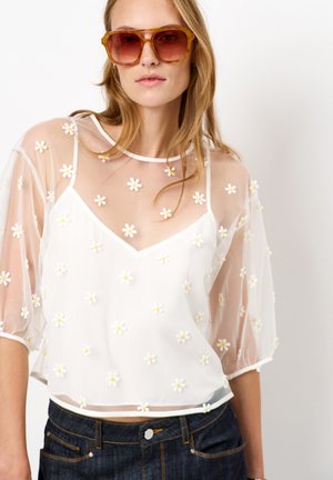 Weiße durchsichtige Bluse mit V-Ausschnitt und kurzen Ballonärmeln, verziert mit kleinen gelben und weißen Blumenapplikationen. Kombiniert mit dunkler Jeans.