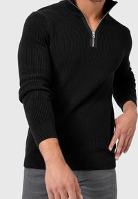 Pull noir côtelé zippé avec un col montant, des manches longues et des fentes latérales, doté d'une fermeture éclair argentée et d'un motif tricot texturé.