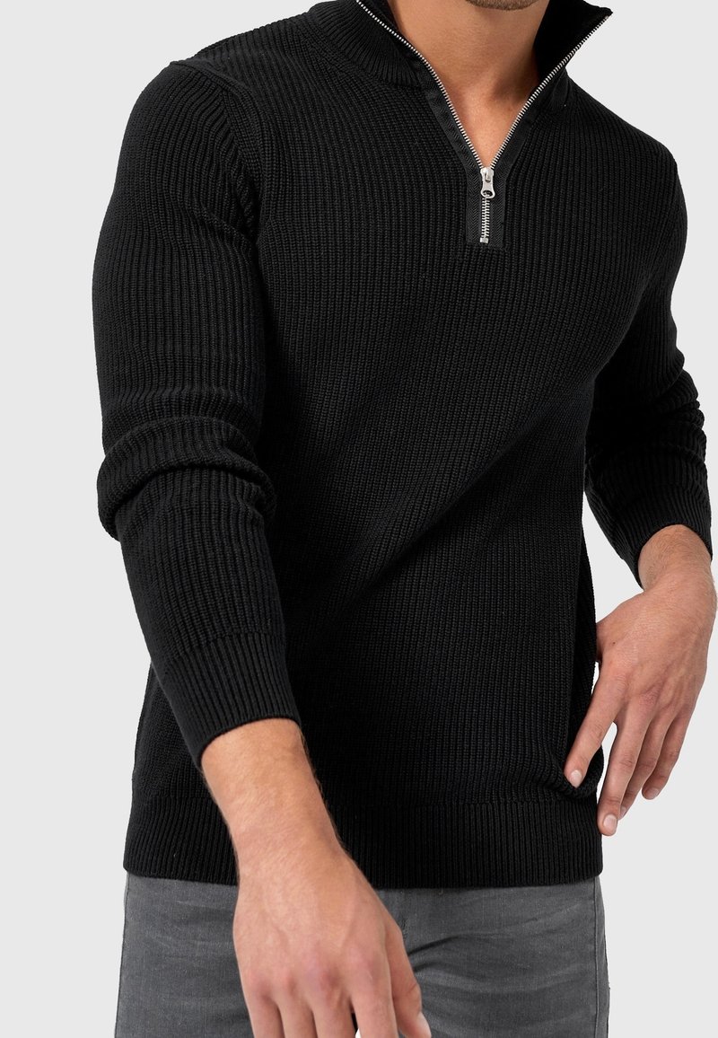 Pull noir côtelé zippé avec un col montant, des manches longues et des fentes latérales, doté d'une fermeture éclair argentée et d'un motif tricot texturé.