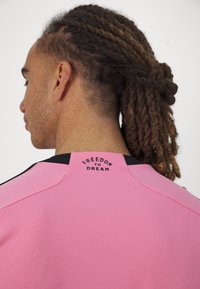 Maglietta sportiva rosa con dettagli neri, con la scritta "LIBERTÀ DI SOGNARE" sul colletto posteriore. Tessuto morbido, design aderente.