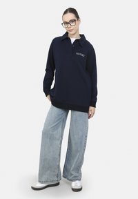 Polo a maniche lunghe di colore blu navy con colletto e testo ricamato. Abbinato a un paio di jeans ampi di colore azzurro chiaro e scarpe da ginnastica bianche.