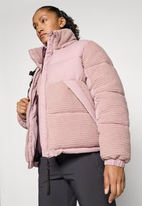 Woman wearing a pink textured puffer jacket with high collar and black pants, looking to the side against a plain light background.