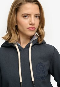 Sudadera con capucha azul marino oscuro con un logo, presenta cordones de color crema y un acabado texturizado. La prenda tiene un cuello en capas con un forro interior colorido.