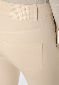 Beige taillierte Hose mit einem strukturierten Taillenband, minimaler Naht und einem Standarddesign für Gesäßtaschen. Glattes Material mit leichtem Glanz.