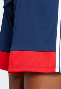 Blauwe sportshorts met een rode ribbelrand, voorzien van witte zijkanten. De stof heeft een gladde textuur, geschikt voor sportactiviteiten.