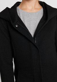 Zwarte hooded jacket van een textuur met ribfluweel, met een ritssluiting en een grijze binnenhemd die eronderuit piekt.