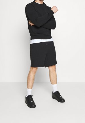 Hombre de pie con los brazos cruzados, vistiendo sudadera negra, camiseta blanca, pantalones cortos negros, calcetines blancos y zapatillas negras, sobre un fondo liso.