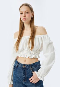 Hvid off-the-shoulder crop top med elastisk volangkant og lange ærmer, parret med højtaljede blå jeans med knaplukning.