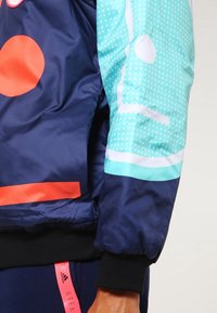 Navy blue bomber jacket featuring a red circle, turquoise patterned sleeve, and elastic cuffs. Material is smooth with a glossy finish.