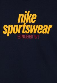 Karaliski zils kokvilnas T-krekls ar izteiktu dzelteno "nike sportswear" tekstu un sarkano "ESTABLISHED 1972" detaļu. Mīksta tekstūra, apaļas kakla dizains.