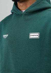 Groene hoodie van zachte stof met een grote capuchon en witte geborduurde logo labels, "STRONGER" en "chance", aan de voorkant.