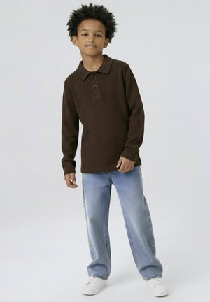 Chlapec s kudrnatými vlasy, oblečený v tmavě hnědém polo s dlouhým rukávem, světle modrých džínách a bílých teniskách stojící před jednoduchým pozadím.