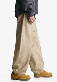 Beige bredde-ben jeans med løs pasform, der har synlige sømme og sidelommer, parret med brune låg- hæl snørebåndsstøvler.