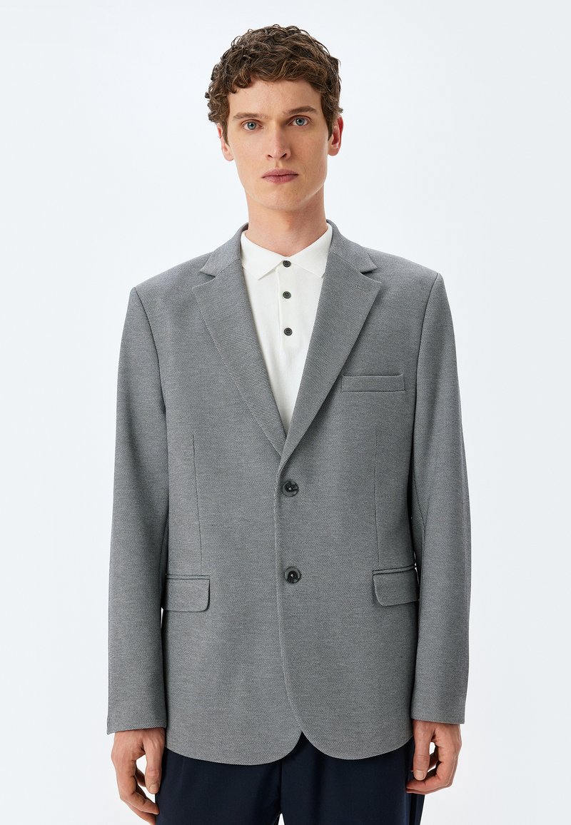 Grau strukturierter Blazer mit Reverskragen, zwei vorderen Knöpfen, seitlichen Taschen und einer Brusttasche, getragen über einem weißen Hemd mit Kragen.