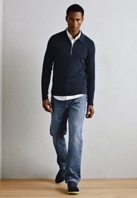 Maglione navy a zip, con camicia bianca sotto, abbinato a jeans denim azzurri chiari e sneaker nere con dettagli verdi.