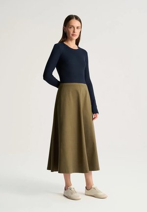 Kobieta stojąca z rękami za plecami, ubrana w granatową bluzkę z długim rękawem, oliwkową spódnicę midi i białe trampki na jednolitym tle.