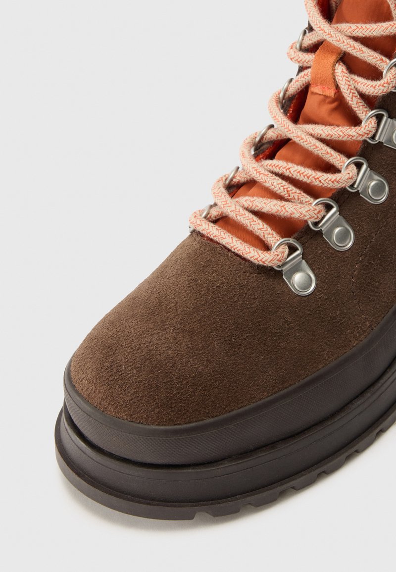 Braune Wildleder-Wanderschuhe mit orangefarbenem Stofffutter, ausgestattet mit silbernen Ösen und beigen Schnürsenkeln. Dicke Gummisohle für guten Halt.