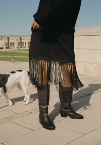 Falda negra con detalles de flecos en el dobladillo, combinada con botas altas negras. El conjunto presenta una tela texturizada y una silueta elegante.