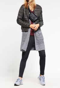 Svart quiltad bomberjacka, grå cardigan, vinröd topp, mörka leggings, grå sneakers och en liten svart crossbody-väska. Texturerade material synliga.