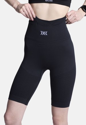 Close-up van een persoon die zwarte naadloze sportshorts met hoge taille en getextureerde panelen en een centraal logo op de tailleband verstelt.