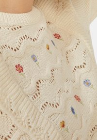 Sweter dzianinowy w kremowym beżu, z haftowanymi kwiatami w kolorze czerwonym, niebieskim i żółtym. Wzór falisty wzdłuż dolnej krawędzi i dekoltu, z detalami w otwartym ściegu.