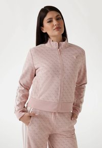 Veste zippée rose clair avec un motif texturé, poignets et taille côtelés. Présente des rayures blanches sur les côtés et un accent logo sur la poitrine.