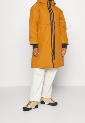 Personne portant un long manteau moutarde avec des détails de fermeture éclair noirs, un pantalon blanc à jambes larges et des baskets beiges avec des semelles noires et des accents orange.