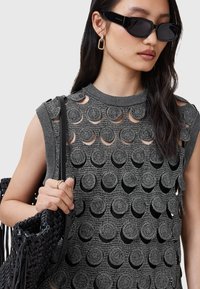 Top gri fără mâneci cu decupaje circulare și tricot texturat. Geantă neagră țesută cu detalii din franjuri. Model purtând ochelari de soare dreptunghiulari negri.