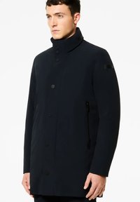 Uomo che indossa un cappotto nero a collo alto con chiusura a bottoni e tasca laterale con zip, rivolto leggermente verso sinistra su uno sfondo bianco uniforme.